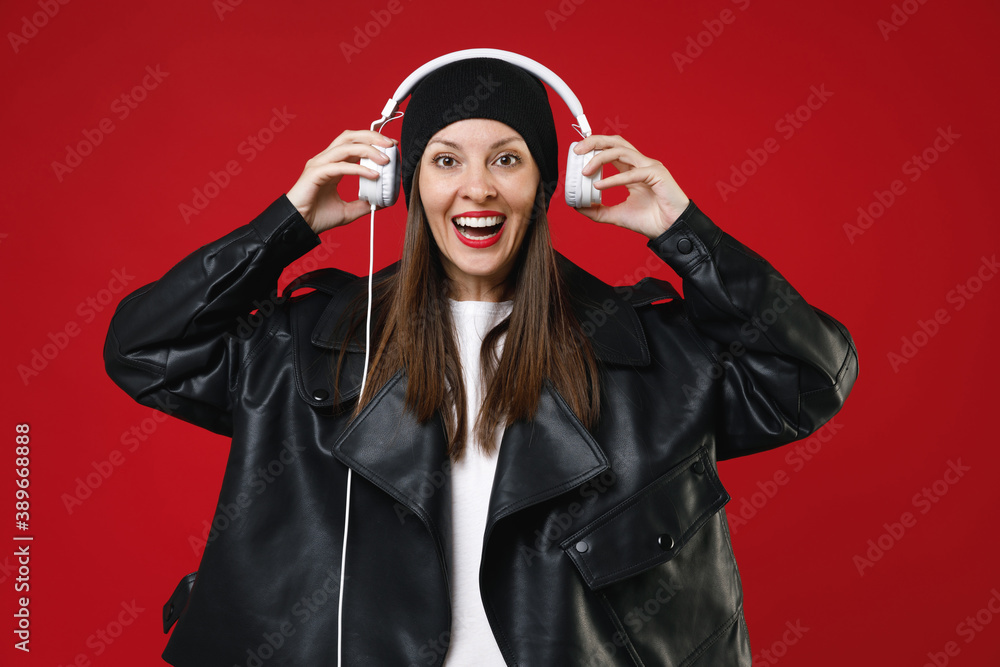 Cheerful funny young brunette woman 20s wearing casual black leather jacket hat standing listening music with headphones looking camera isolated on bright red colour wall background studio portrait. Cheerful funny young brunette woman 20s wearing casual black leather jacket hat standing listening music with headphones looking camera isolated on bright red colour wall background studio portrait.