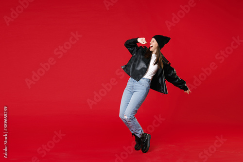 Full length of funny cheerful young brunette woman 20s in casual black leather jacket white t-shirt hat standing on toes dancing sing song isolated on bright red colour background studio portrait.
