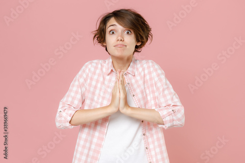Pleading begging young brunette woman 20s wearing casual basic checkered shirt standing holding hands folded in prayer looking camera isolated on pastel pink colour wall background, studio portrait.