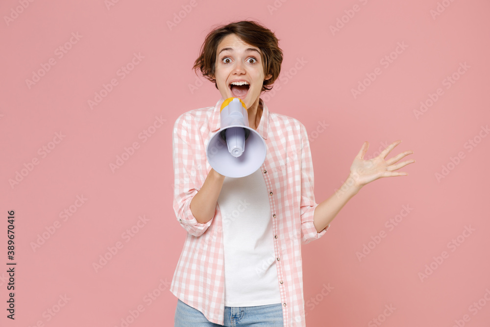Excited surprised shocked amazed young brunette woman 20s wearing casual checkered shirt standing screaming in megaphone spreading hands isolated on pastel pink colour background, studio portrait. Excited surprised shocked amazed young brunette woman 20s wearing casual checkered shirt standing screaming in megaphone spreading hands isolated on pastel pink colour background, studio portrait.