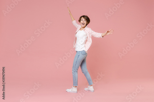 Full length side view of excited surprised young brunette woman 20s wearing casual checkered shirt standing spreading hands looking camera isolated on pastel pink colour background, studio portrait.