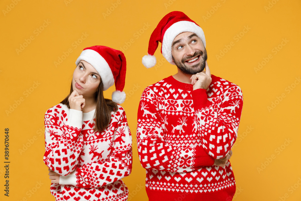 Fototapeta premium Pensive young Santa couple friends man woman 20s in red sweater Christmas hat put hand prop up on chin isolated on yellow background studio portrait. Happy New Year celebration merry holiday concept.