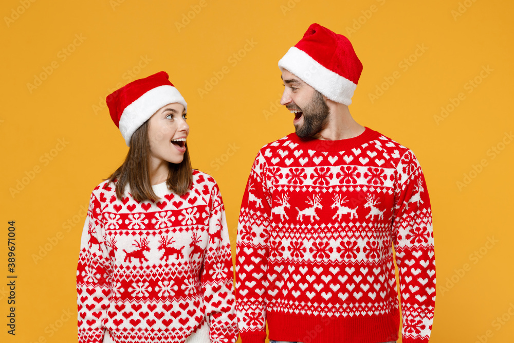 Excited young Santa couple friends man woman in red sweater, Christmas hat keeping mouth open looking at each other isolated on yellow background studio. Happy New Year celebration holiday concept.