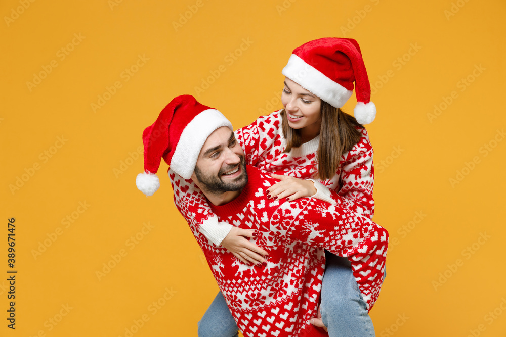 Smiling young Santa couple friends man woman in sweater Christmas hat giving piggyback ride to joyful, sit on back looking at each other isolated on yellow background. Happy New Year holiday concept. Smiling young Santa couple friends man woman in sweater Christmas hat giving piggyback ride to joyful, sit on back looking at each other isolated on yellow background. Happy New Year holiday concept.