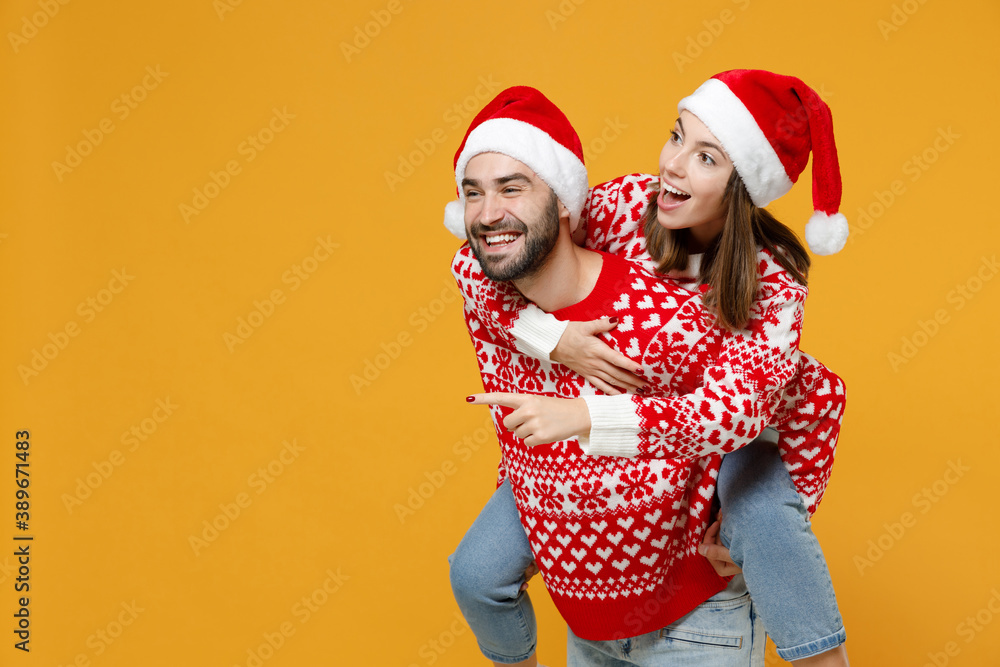 Fototapeta premium Excited young Santa couple friends man woman in Christmas hat giving piggyback ride to joyful, sit on back pointing index finger aside isolated on yellow background. Happy New Year holiday concept.
