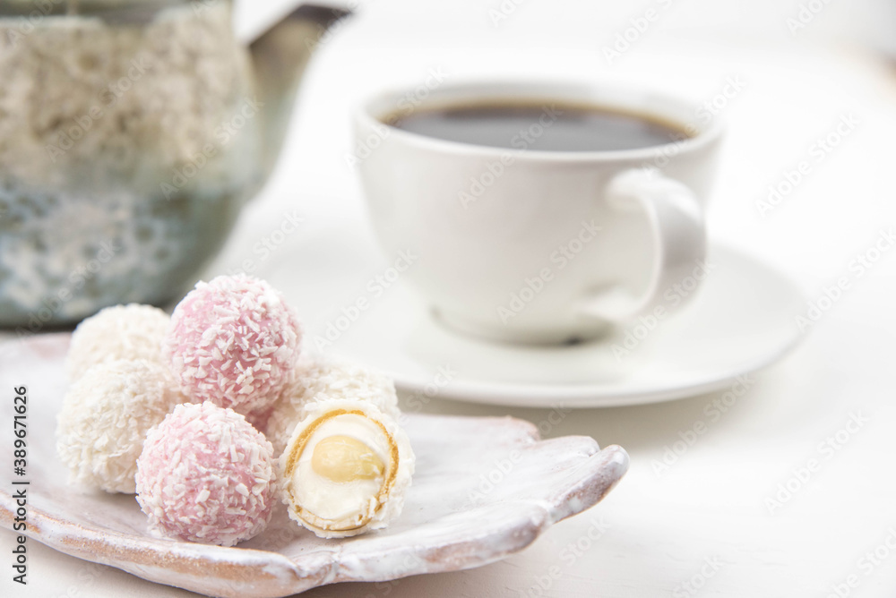 White coconut candy balls on handmade plate. Coconut cookies on white background with the cup of tea. Teapot with black tea. White coconut candy balls on handmade plate. Coconut cookies on white background with the cup of tea. Teapot with black tea.