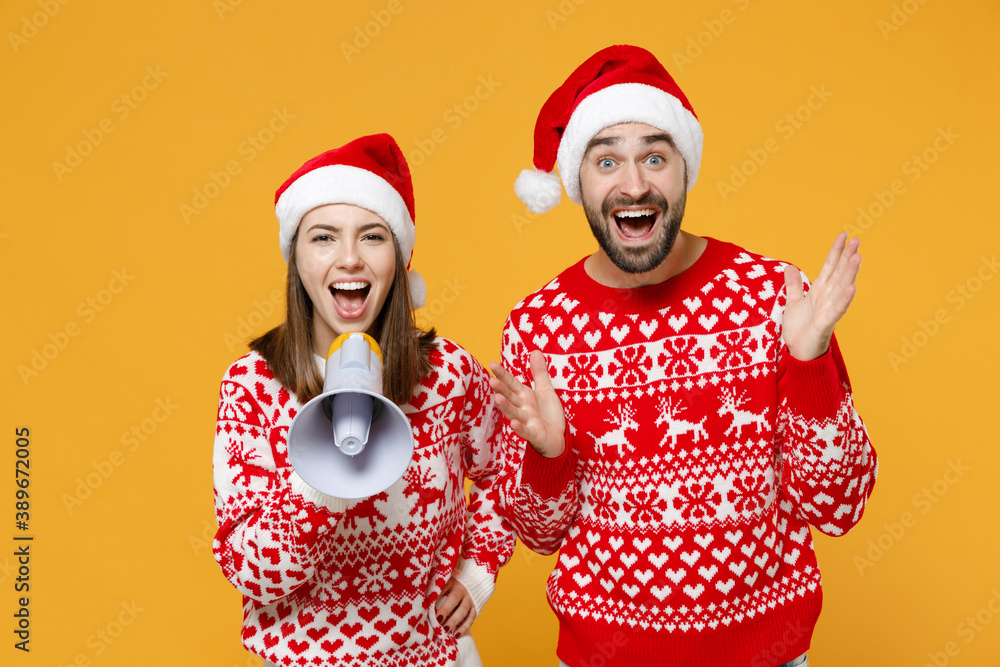Surprised young Santa couple friends man woman in sweater Christmas hat screaming in megaphone spreading hands isolated on yellow background studio. Happy New Year celebration merry holiday concept. Surprised young Santa couple friends man woman in sweater Christmas hat screaming in megaphone spreading hands isolated on yellow background studio. Happy New Year celebration merry holiday concept.
