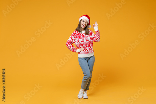 Full length of blinking young Santa woman 20s wearing red sweater, Christmas hat showing OK gesture isolated on yellow background, studio portrait. Happy New Year celebration merry holiday concept.