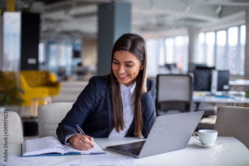 Young businesswoman working on the laptop, writing notes in her notebook in the workplace.