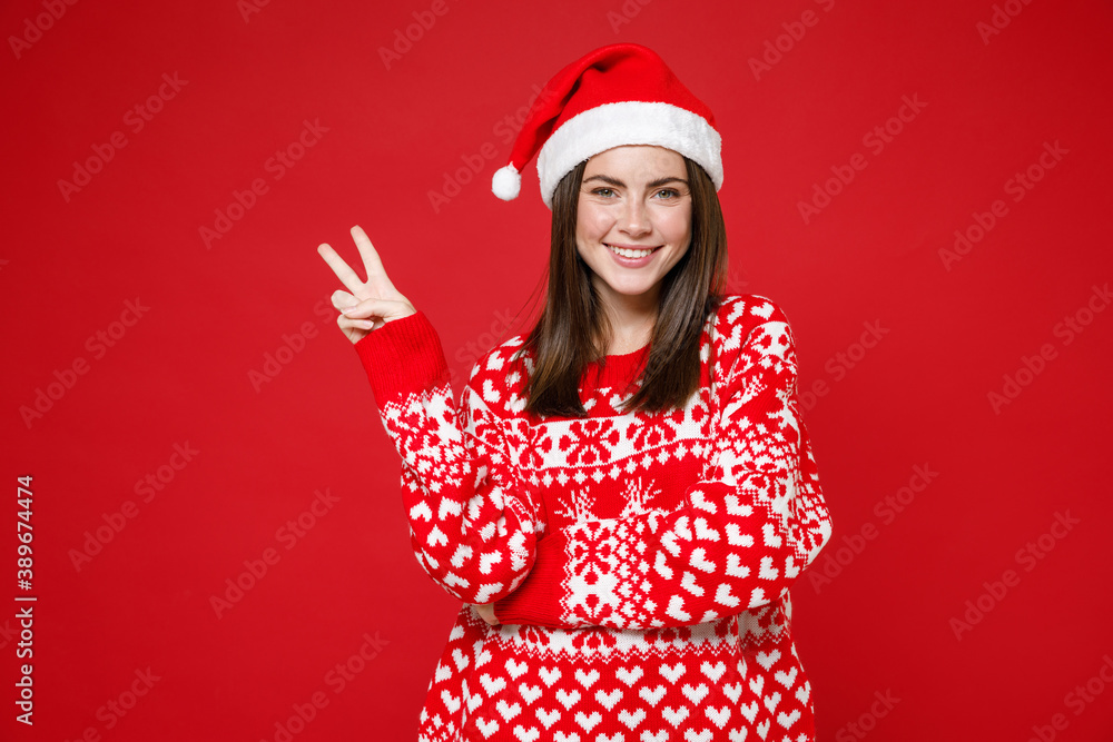 Smiling funny young brunette Santa woman 20s wearing sweater Christmas hat showing victory sign isolated on red colour background, studio portrait. Happy New Year celebration merry holiday concept. Smiling funny young brunette Santa woman 20s wearing sweater Christmas hat showing victory sign isolated on red colour background, studio portrait. Happy New Year celebration merry holiday concept.