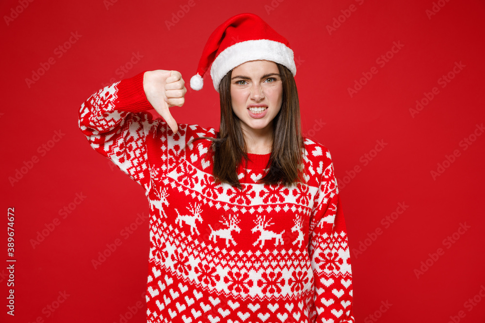 Dissatisfied young brunette Santa woman 20s in sweater, Christmas hat showing thumb down isolated on bright red colour background, studio portrait. Happy New Year celebration merry holiday concept. Dissatisfied young brunette Santa woman 20s in sweater, Christmas hat showing thumb down isolated on bright red colour background, studio portrait. Happy New Year celebration merry holiday concept.
