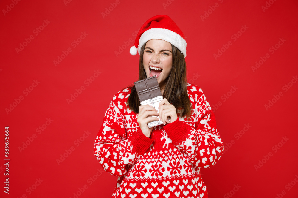 Cheerful crazy young Santa woman 20s wearing sweater Christmas hat biting eating chocolate bar isolated on bright red wall background studio portrait. Happy New Year celebration merry holiday concept. Cheerful crazy young Santa woman 20s wearing sweater Christmas hat biting eating chocolate bar isolated on bright red wall background studio portrait. Happy New Year celebration merry holiday concept.