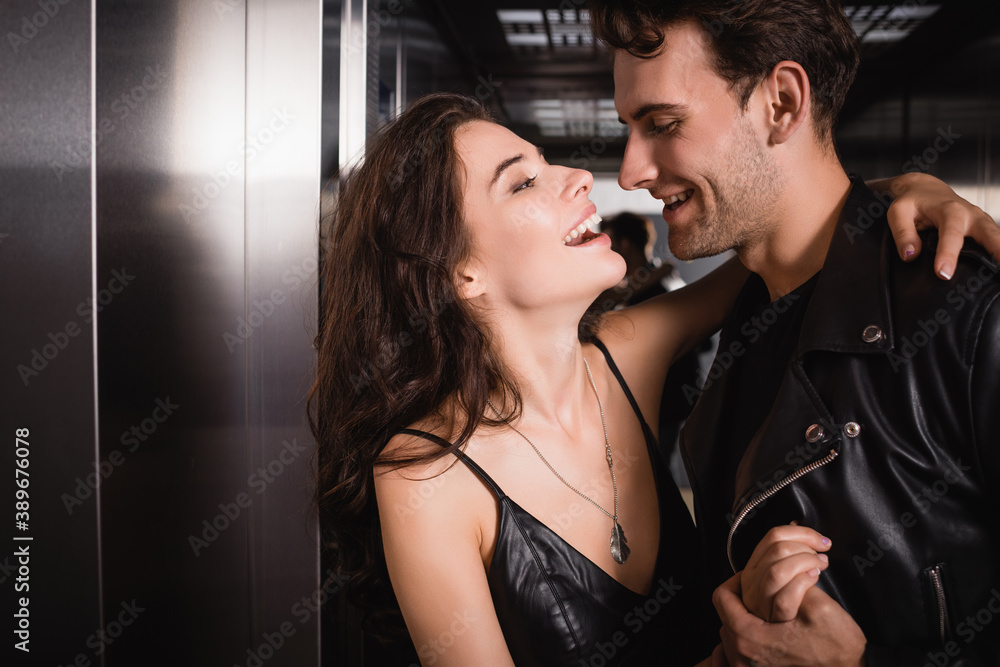 Brunette woman laughing, while embracing and looking at smiling man in leather jacket in elevator Brunette woman laughing, while embracing and looking at smiling man in leather jacket in elevator