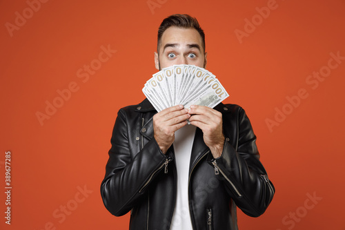 Obraz na plátně Shocked young bearded man in basic white t-shirt, black leather jacket standing covering face with fan of cash money in dollar banknotes looking camera isolated on orange background studio portrait