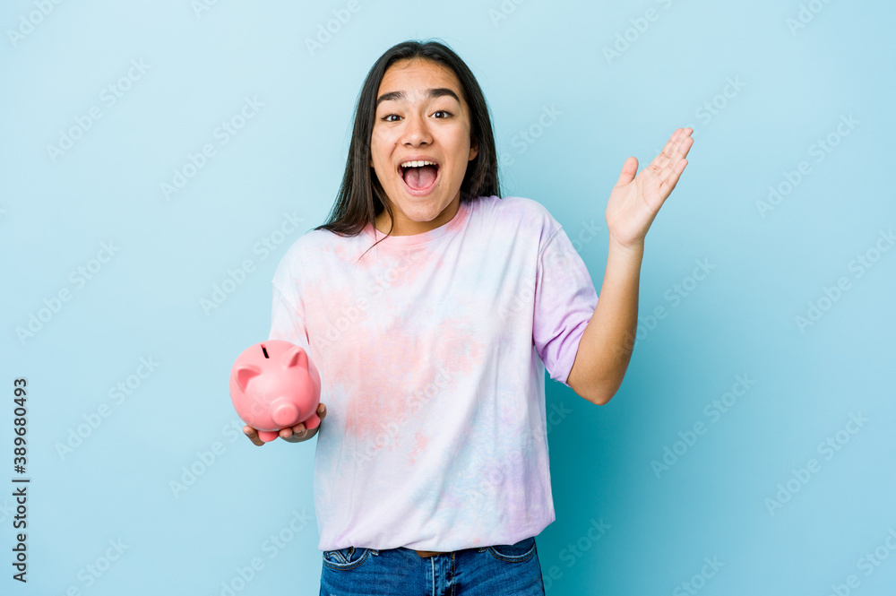 Young asian woman holding a pink bank over isolated background receiving a pleasant surprise, excited and raising hands. Young asian woman holding a pink bank over isolated background receiving a pleasant surprise, excited and raising hands.