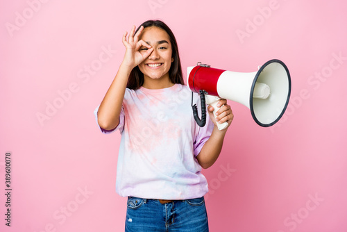 Young asian woman holding a megaphone isolated on pink background excited keeping ok gesture on eye.