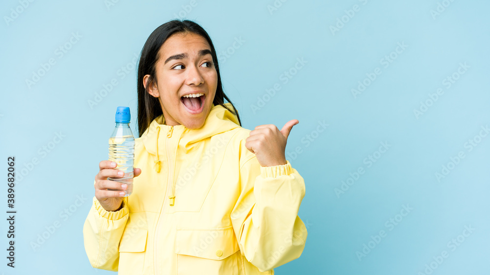 Young asian woman holding a bottle of water isolated on blue background points with thumb finger away, laughing and carefree. Young asian woman holding a bottle of water isolated on blue background points with thumb finger away, laughing and carefree.