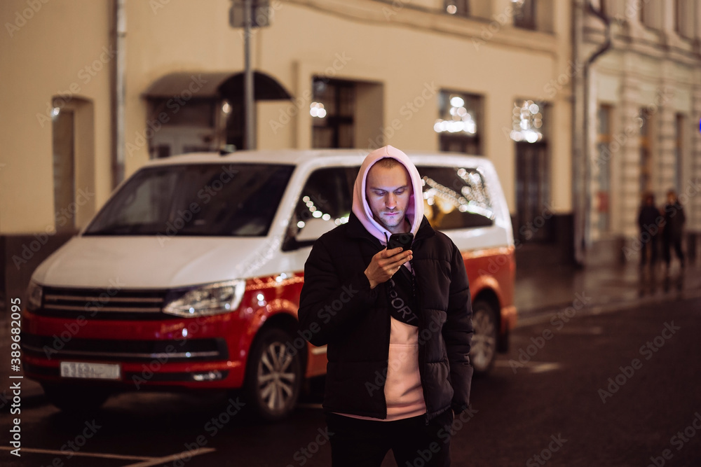 Man in hood looking into his smartphone in background of car on night street.