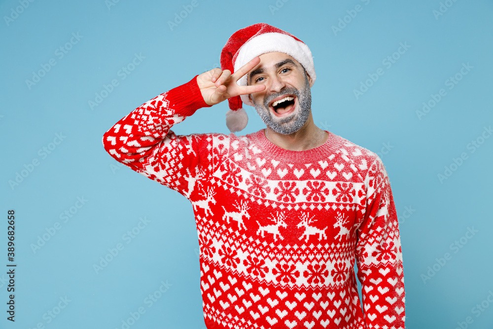Excited young Northern bearded man frozen icy snow face in Santa hat Christmas sweater showing victory sign isolated on blue background. Happy New Year celebration merry holiday winter time concept. Excited young Northern bearded man frozen icy snow face in Santa hat Christmas sweater showing victory sign isolated on blue background. Happy New Year celebration merry holiday winter time concept.