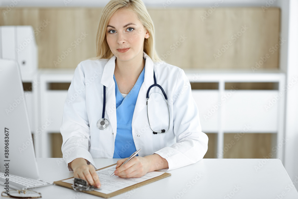 Portrate of woman-doctor at work while sitting at the desk in clinic. Blonde cheerful physician filling up medical form or prescription Portrate of woman-doctor at work while sitting at the desk in clinic. Blonde cheerful physician filling up medical form or prescription