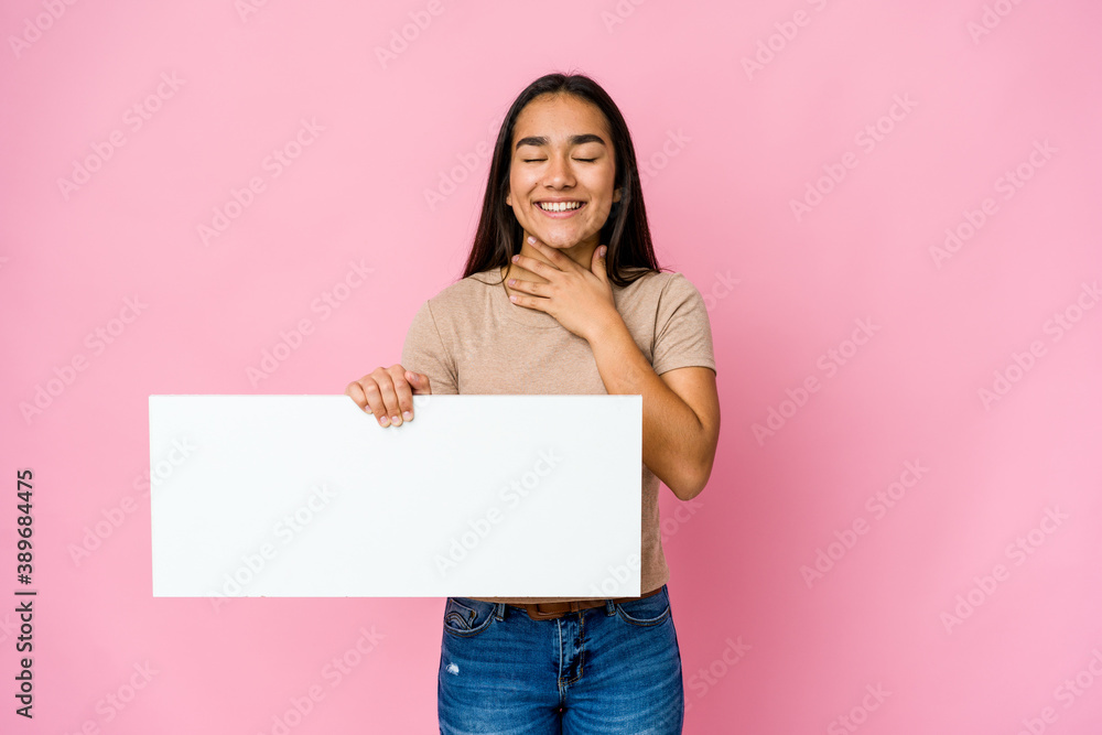 Young asian woman holding a blank paper for white something over isolated background laughs out loudly keeping hand on chest. Young asian woman holding a blank paper for white something over isolated background laughs out loudly keeping hand on chest.