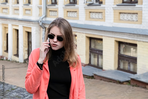 Young stylish woman in coral jacket and sunglasses is displeased talking on a mobile phone while walking down the street