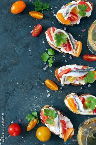 Bruschetta with light cheese, tomato and anchovy served with white wine on dark background. Top view with copy space.