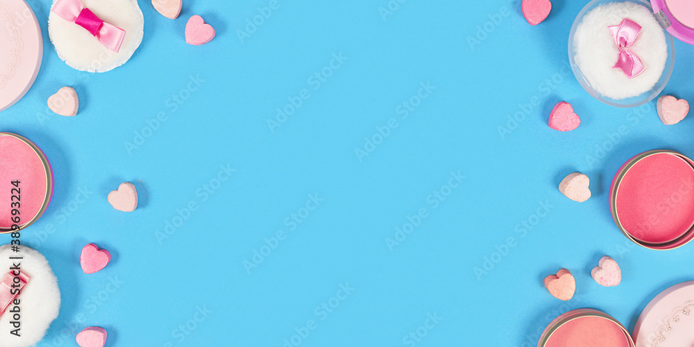 Banner with pink blush beauty products and powder puffs with ribbons and heart shaped pressed powder framing blue background with empty copy space