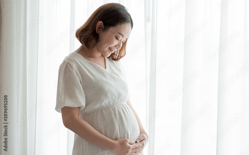 Asian pregnant woman wearing white dress and standing beside window Asian pregnant woman wearing white dress and standing beside window