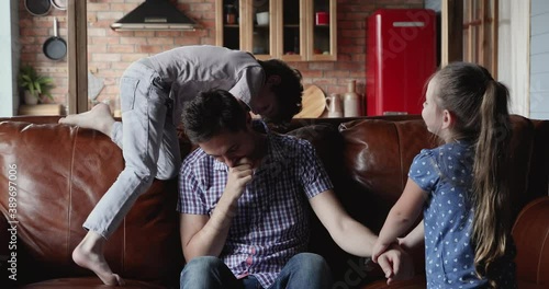 Noisy active kids want to play with daddy sluggish dad sitting on sofa feels exhausted to spend time with children. Hyperactive son and daughter, upbringing problems lack of energy need repose concept