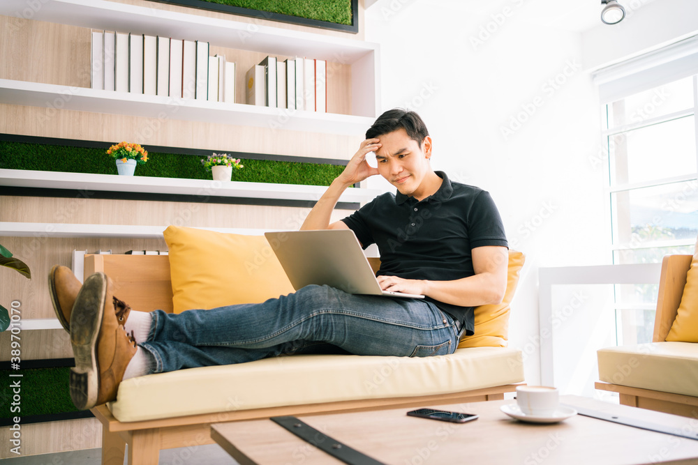 Asian male in casual feels stress and concerned with laptop while working and sitting on the sofa. Businessman working with problems on a laptop at home. Solving the problem process in business.