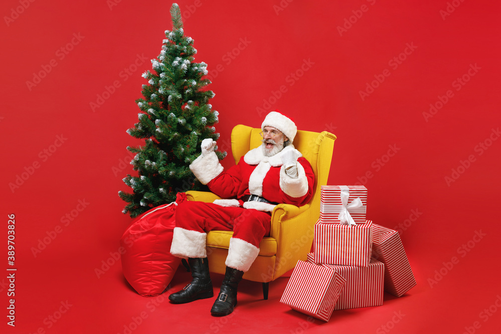 Excited Santa Claus man in Christmas hat suit coat sit in armchair with fir tree present gifts doing winner gesture isolated on red background studio. Happy New Year celebration merry holiday concept.