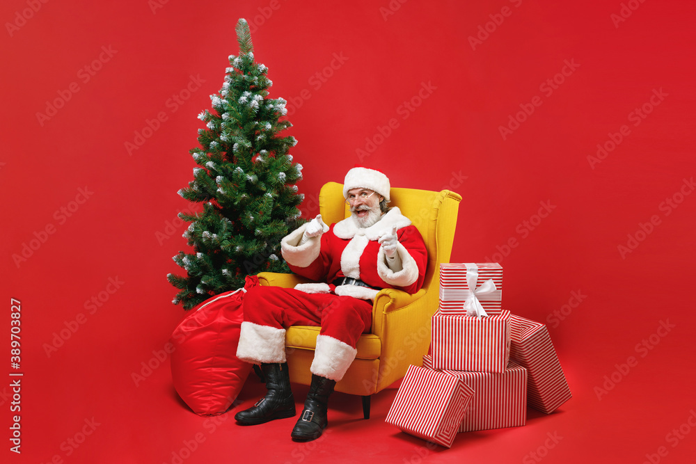 Excited Santa Claus man in Christmas hat suit coat sit in armchair with fir tree presents gifts pointing index fingers on camera isolated on red background. Happy New Year celebration holiday concept. Excited Santa Claus man in Christmas hat suit coat sit in armchair with fir tree presents gifts pointing index fingers on camera isolated on red background. Happy New Year celebration holiday concept.