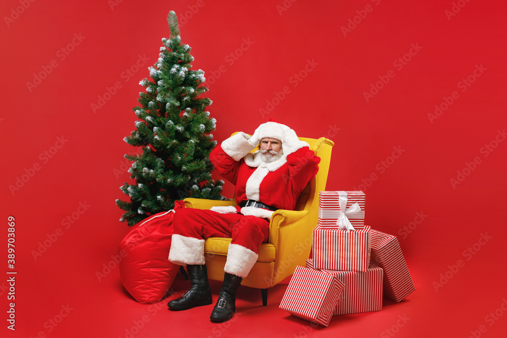 Displeased Santa Claus man in Christmas hat suit sit in armchair with fir tree presents gifts put hands on head isolated on red background studio. Happy New Year celebration merry holiday concept.