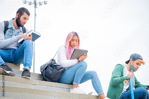 Young people university studyng outoor during coronavirus pandemic. Three multietnic university students with protective masks sitting outdoor on the stairs keeping the social distance photo