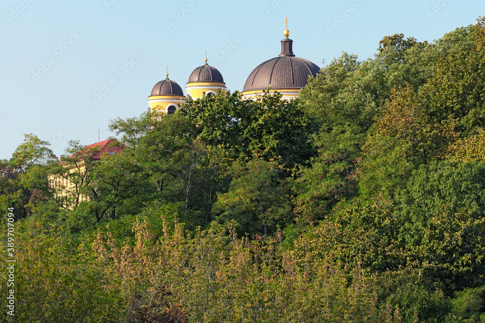 The three domes of an ancient church hidden in a green leaves of trees against blue sky. The Cathedral of St. Alexander of Kyiv. Autumn morning landscape. Kyiv, Ukraine