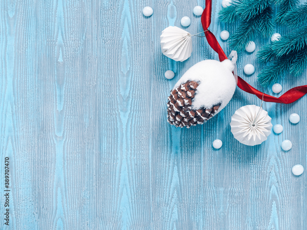 Fototapeta premium Christmas composition. A large decorative pine cone and paper decorations are on a blue wooden background. Coniferous branches and a red ribbon are nearby. Flat lay, top view, copy space.