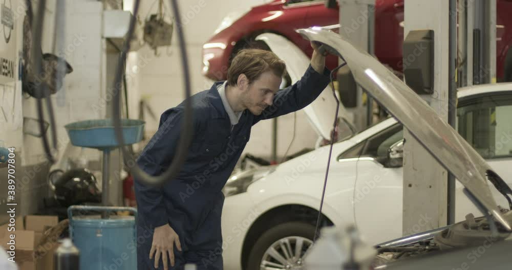 Male car mechanic looking under vehicle bonnet in automotive vehicle repair workshop Male car mechanic looking under vehicle bonnet in automotive vehicle repair workshop