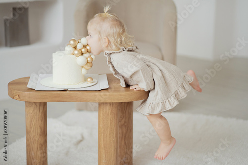 little blonde girl eats her sweet cream cake with balloons for her birthday. Dressed in a light beige stylish dress and goes barefoot, celebrates the holiday