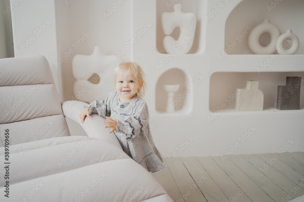 little blonde girl celebrating her birthday playing on the couch, running around having fun in a comfortable baby dress. bright and stylish interior of the new apartment. little blonde girl celebrating her birthday playing on the couch, running around having fun in a comfortable baby dress. bright and stylish interior of the new apartment.