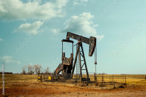 Rusty oil well pump jack sitting in winter pasture with rough hay stubble and clay covered ground and bare trees on horizon under pretty blue sky