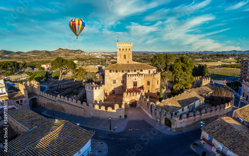 Aerial view of historic Benisano castle near Valencia Spain with loopholes, towers, walls colorful hot air balloon flying by, dreamy blue sky