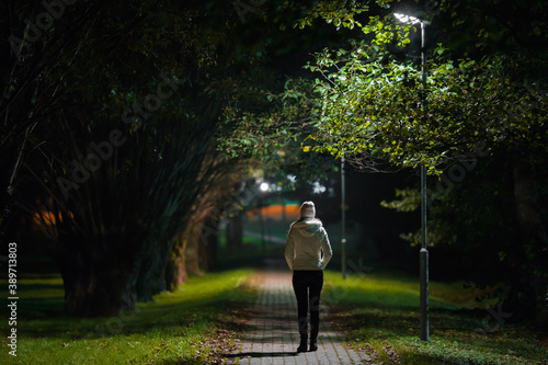 One young adult woman in white jacket walking on sidewalk through alley of trees under lamp light in autumn night. Spending time alone in nature. Peaceful atmosphere. Back view. photo