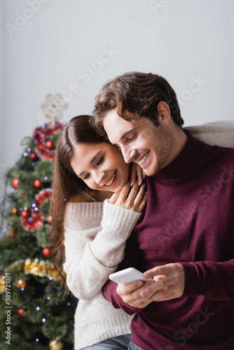 happy couple in sweaters looking at smartphone near christmas tree on blurred background