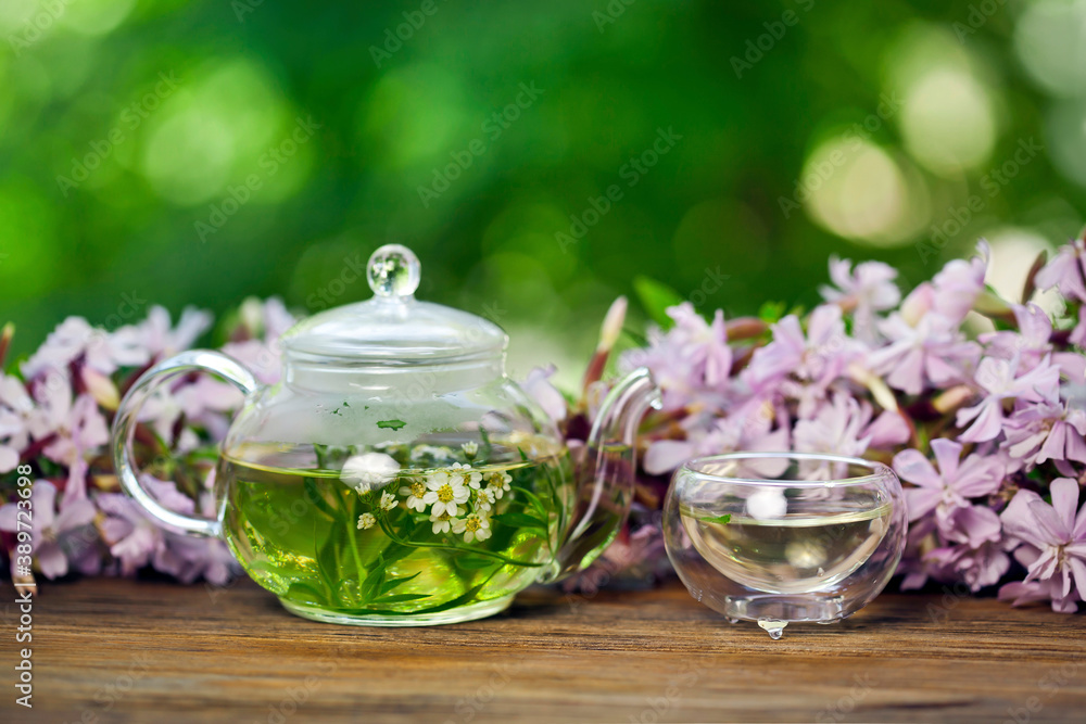 Herbal tea in a glass cup and teapot with bouquet of pink flowers on a green bokeh background. Summer tea ceremony in a garden. Herbal tea in a glass cup and teapot with bouquet of pink flowers on a green bokeh background. Summer tea ceremony in a garden.