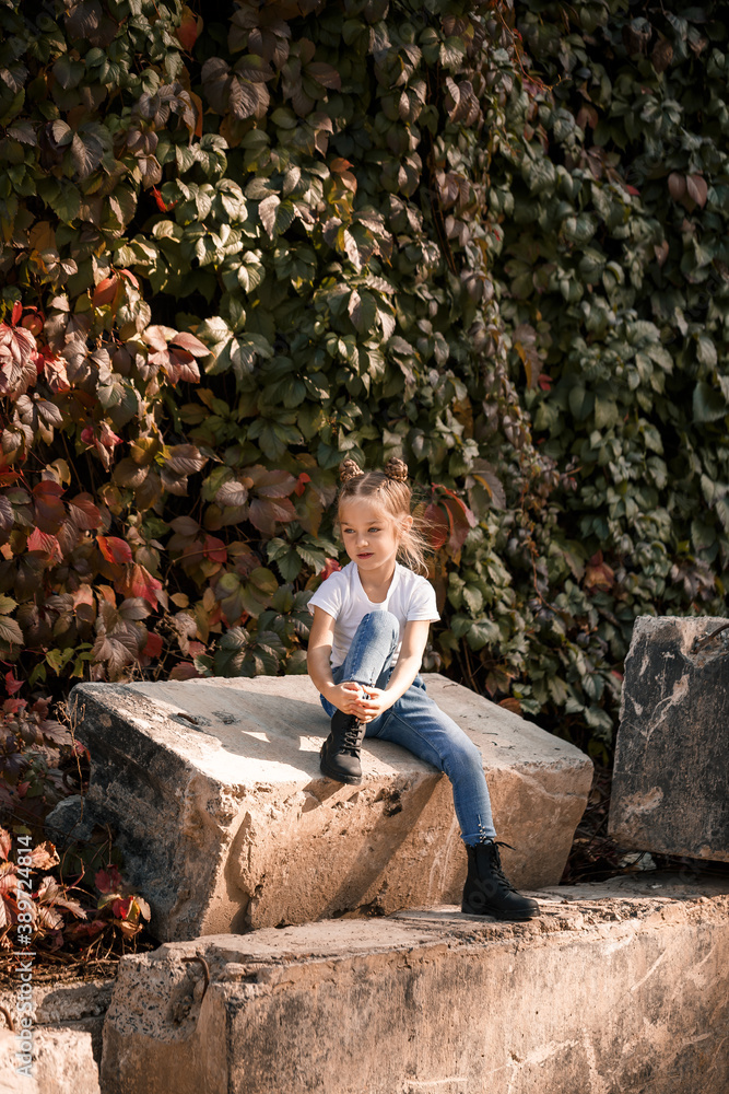 Street photo of a little beautiful girl in jeans and a white t-shirt on a background of concrete slabs and autumn leaves Street photo of a little beautiful girl in jeans and a white t-shirt on a background of concrete slabs and autumn leaves