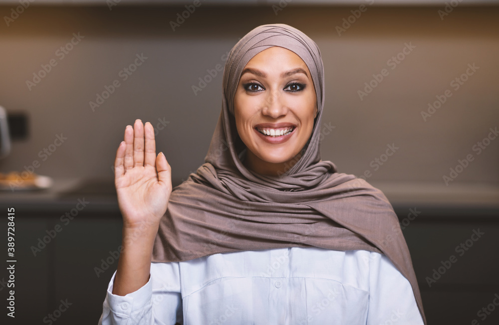 Muslim Woman Waving Hello Smiling To Camera Sitting In Kitchen