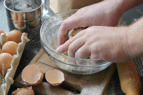 Hands kneading dough. Ball of dough on a rustic wooden background with dusting of flour. Dough with flour near broken egg with yolk in bowl and other utensil, ingredients lies on dark concrete table. 