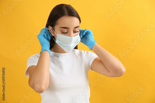 Woman in medical gloves putting on protective face mask against yellow background