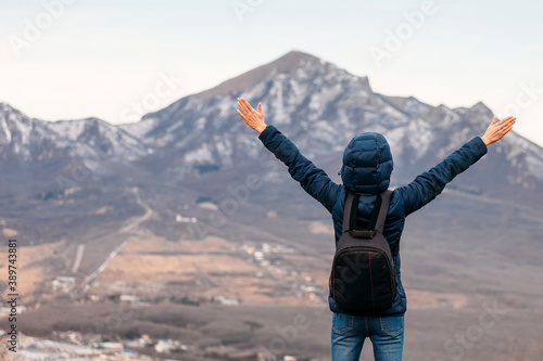 Young woman in a jacket and a backpack on the top of the mountain with arms raised up